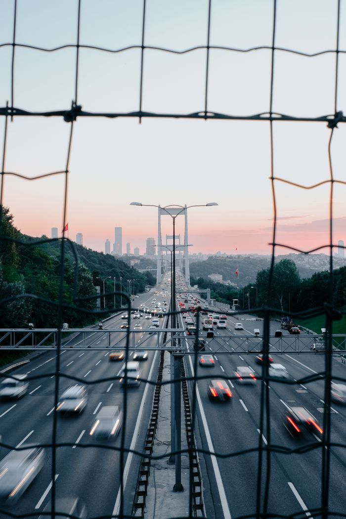 Home Blurred cars moving swiftly on a highway with a cityscape background, captured at dusk through a fence.